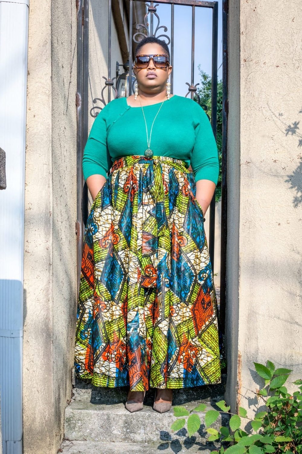 A woman standing in a doorway in a green top and a printed skirt, looking directly at the camera.